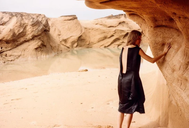Woman in black satin dress poses near rocky landscape with water, creating a striking fashion and nature-themed visual narrative.