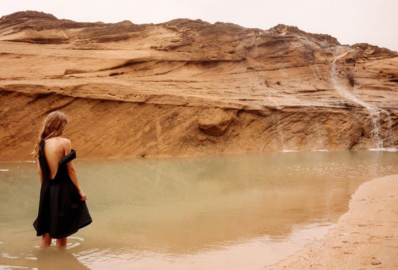 Woman in a black dress standing in shallow water near a textured rock formation with a small waterfall in a desert landscape.