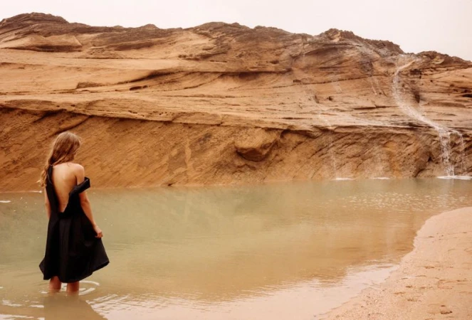 Woman in a black dress standing in shallow water near a textured rock formation with a small waterfall in a desert landscape.