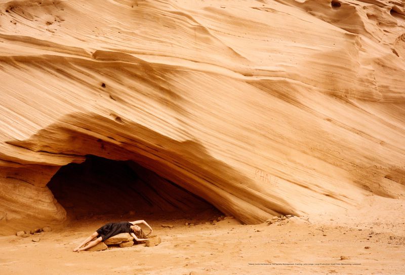Woman in black dress lays on rocks near cave entrance in desert landscape with layered sandstone formations, ochre tones and textural patterns.
