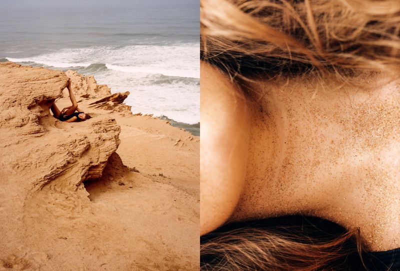 Woman in black swimsuit lies in rock formation overlooking ocean with waves; detailed close up of skin and hair covered in sand. 
