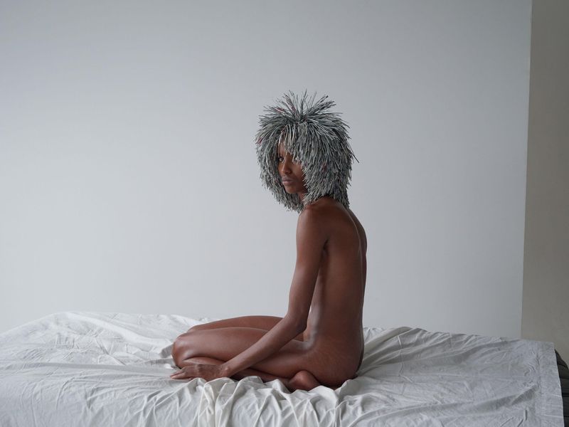 Nude adult female with grey, spiked headpiece sits on a white bed against a plain background in a portrait shot with soft, diffused lighting.