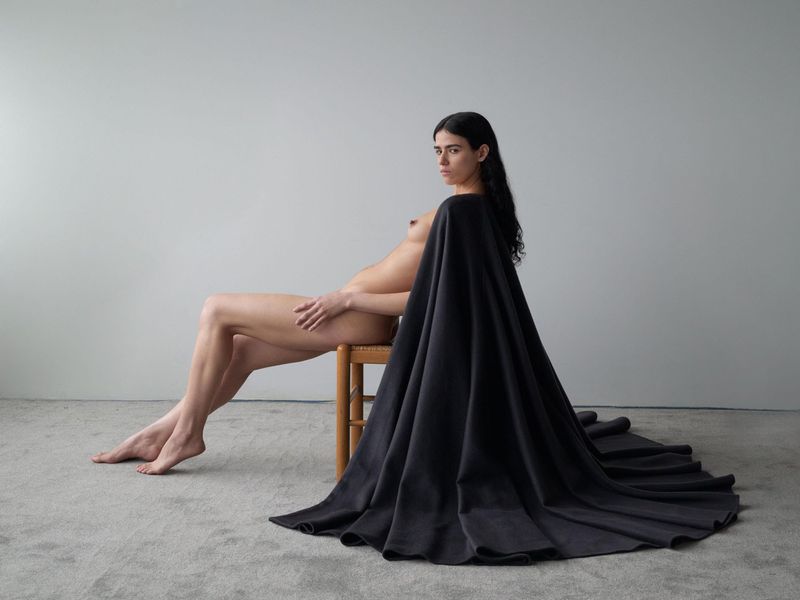 Nude woman with long dark hair sits on a wooden chair in a minimalist room draped with a dark fabric, looking directly at the camera.