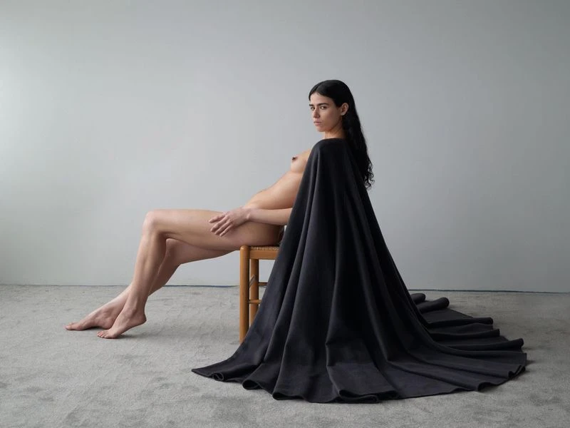 Nude woman with long dark hair sits on a wooden chair in a minimalist room draped with a dark fabric, looking directly at the camera.