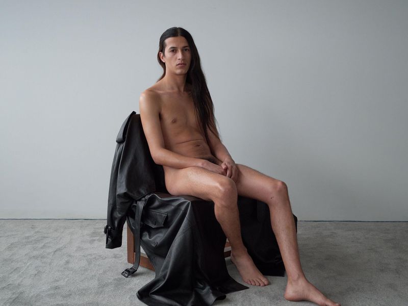 Nude male model sits on chair in photo studio with long dark hair and black leather jacket. Light gray seamless backdrop and floor.