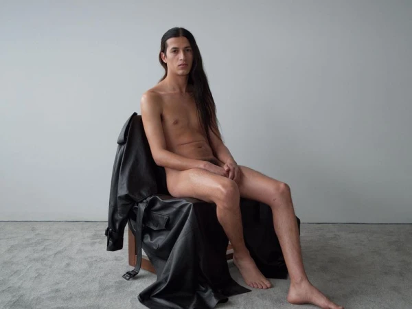 Nude male model sits on chair in photo studio with long dark hair and black leather jacket. Light gray seamless backdrop and floor.