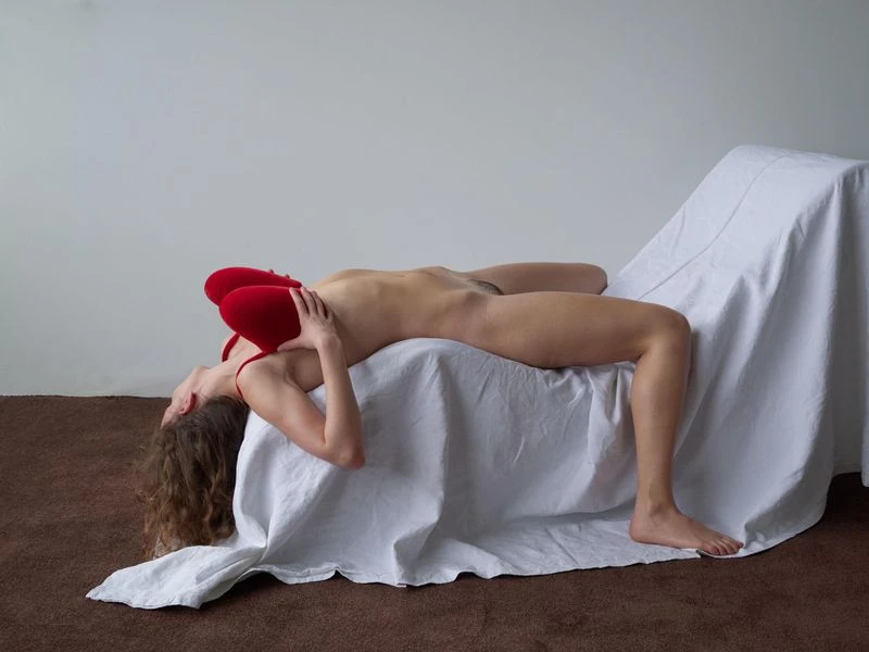 A nude adult female reclines on a draped surface with a red top and bright red earmuffs, against a plain white backdrop with a brown floor.