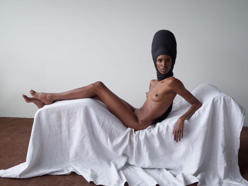 Woman posing nude on a white sheet, wearing a black head covering, sitting indoors on a brown floor against a plain white wall.