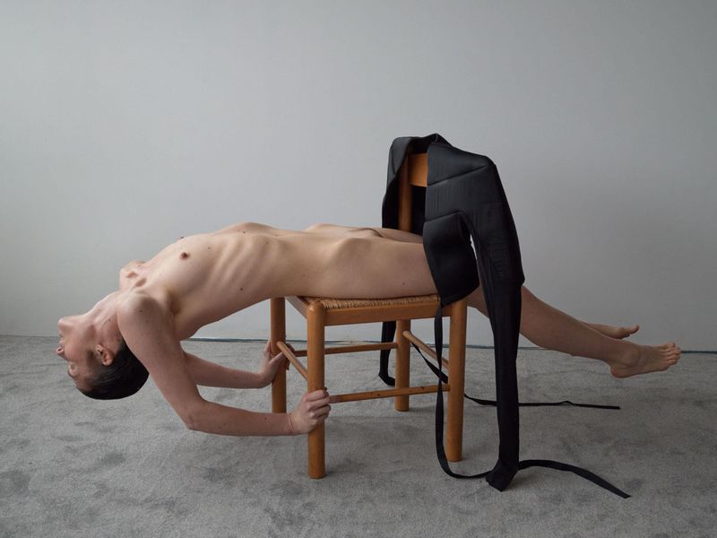 Artistic nude full-body shot of an adult female lying across a light wooden chair against a grey backdrop with black cloth draped.