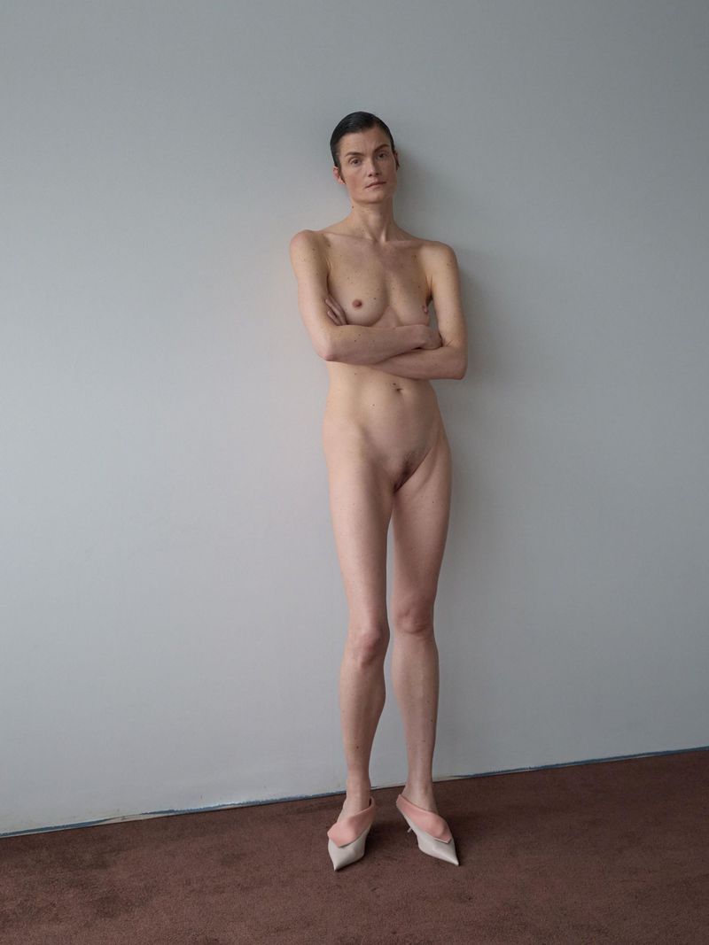 Full-body portrait of a slender adult woman with arms crossed, standing nude in heels against a neutral wall with a brown carpet.