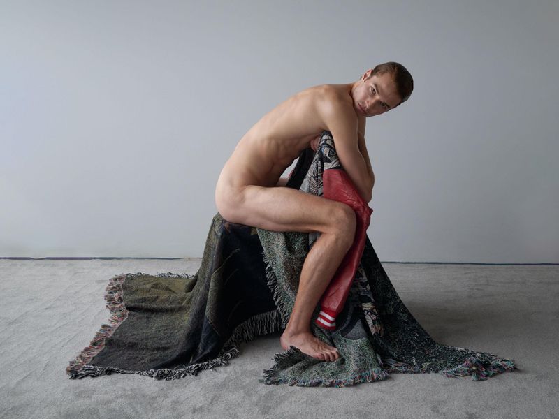 Adult male posing indoors, seated on draped, patterned blanket with neutral wall. He is shirtless, looking at the camera.