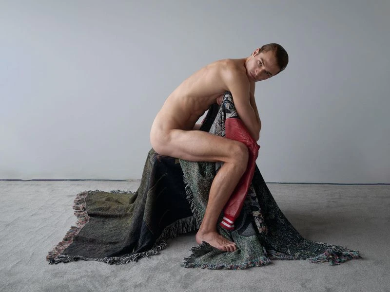 Adult male posing indoors, seated on draped, patterned blanket with neutral wall. He is shirtless, looking at the camera.