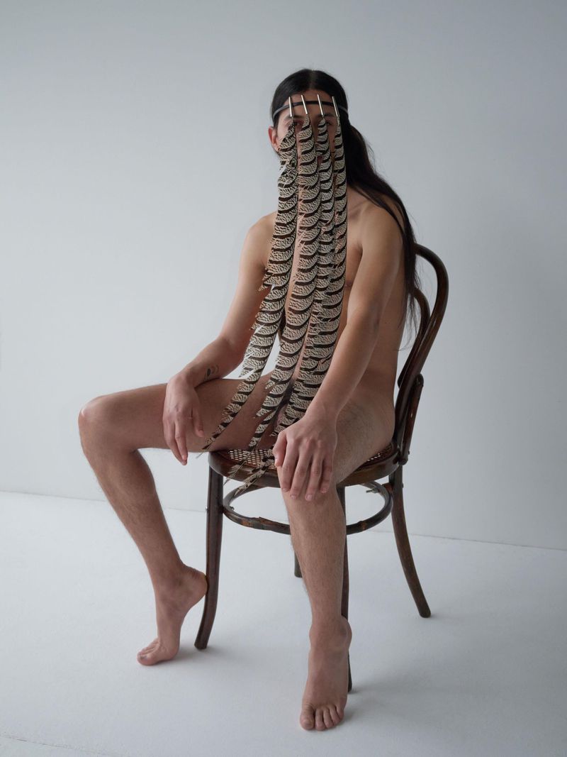 Person with long dark hair seated on a wooden chair, wearing a headband hung with patterned feathers covering their face.