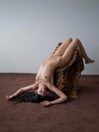 Nude woman with dark curly hair reclines on animal print fabric, indoors with a gray wall and brown floor, in a studio setting.