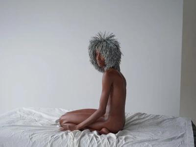 Model with textured grey spiky hair kneeling on a white draped surface against a plain white wall