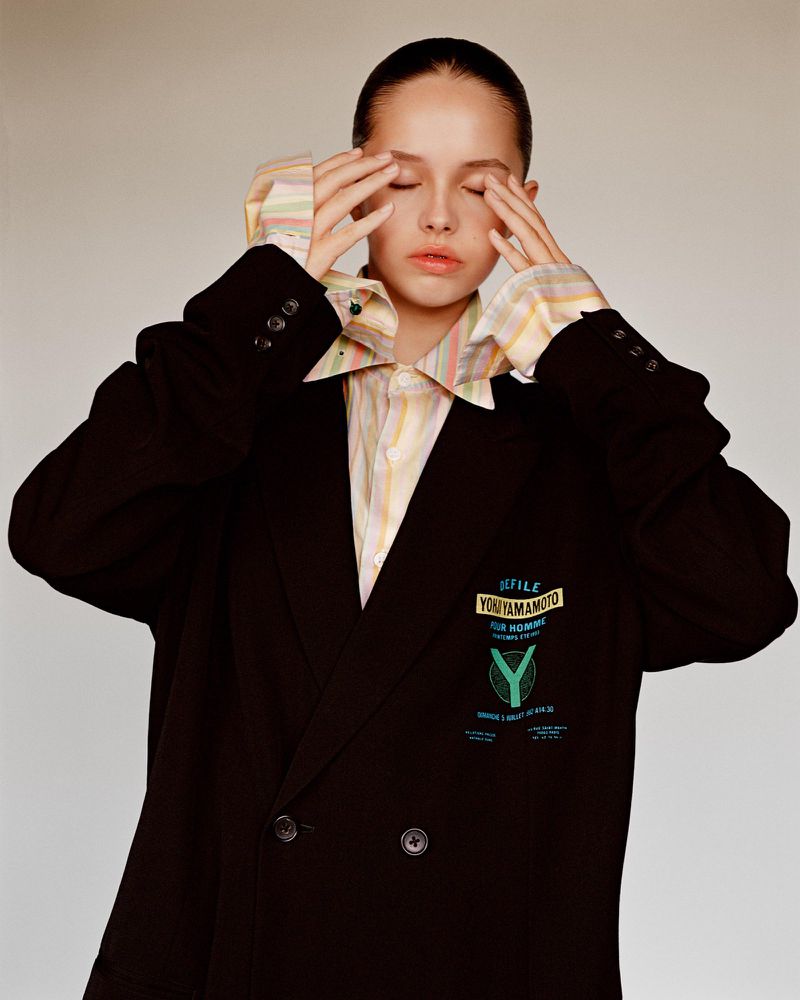 Portrait of a young woman with slicked-back hair wearing an oversized black blazer with hands near her face, eyes closed, and neutral expression.