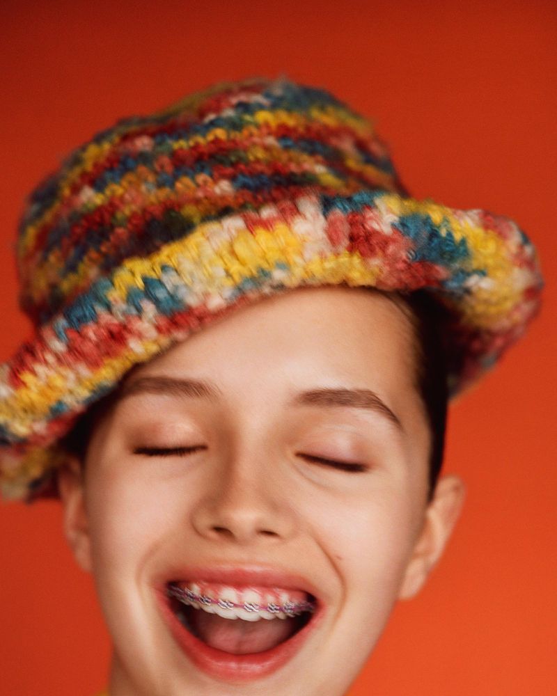 Smiling young person with braces, eyes closed, and wearing a colorful knit hat, set against a solid orange backdrop in a close-up shot.