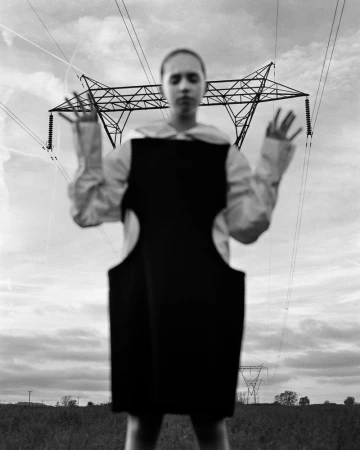 Monochrome portrait of a blurred figure in a black dress posing outdoors against a backdrop of power lines and a cloudy sky.