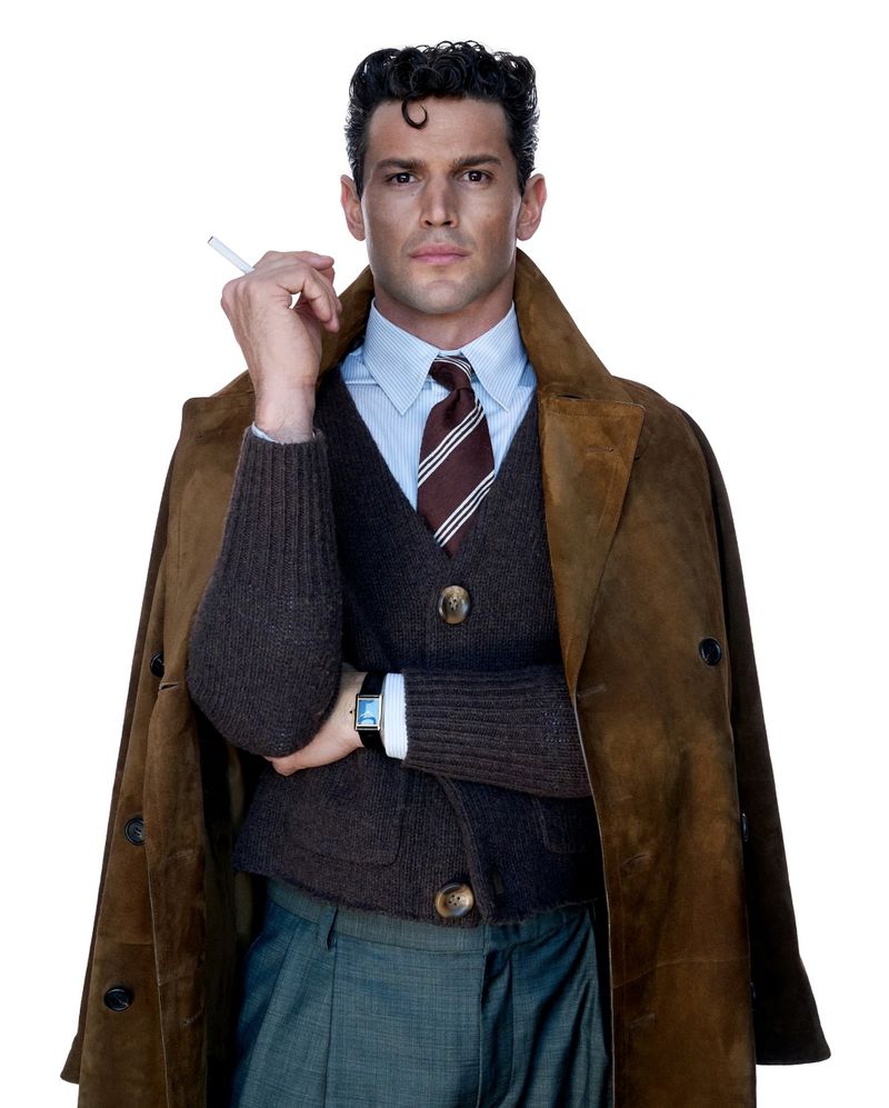 Man wearing a brown suede coat over a dark knit vest, striped tie, and holding a cigarette against a plain white background.
