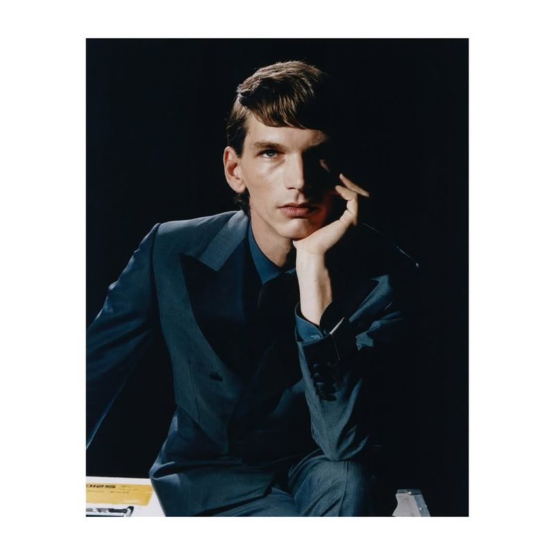 Portrait of a young adult male in a blue suit, resting his head on his hand, against a dark backdrop with subtle lighting, creating a sophisticated mood.