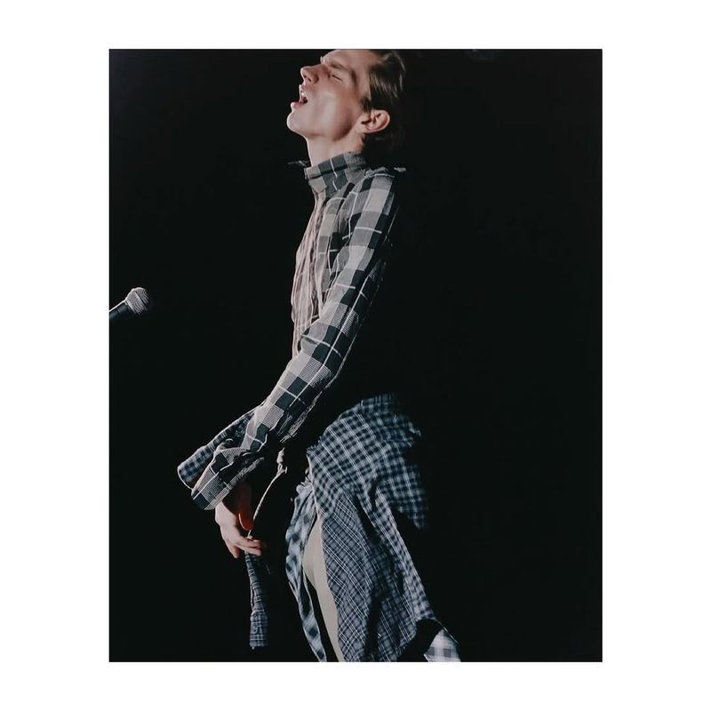 Male singer with a plaid shirt and open mouth performing in front of a microphone against a dark background on stage.
