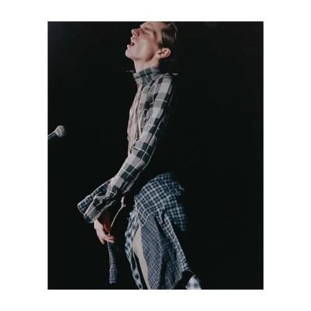 Male singer with a plaid shirt and open mouth performing in front of a microphone against a dark background on stage.