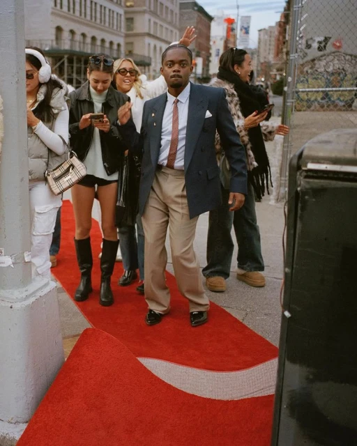 A man walks on a red carpet on a city sidewalk surrounded by a small crowd, captured in an eye-level medium shot on a sunny day.