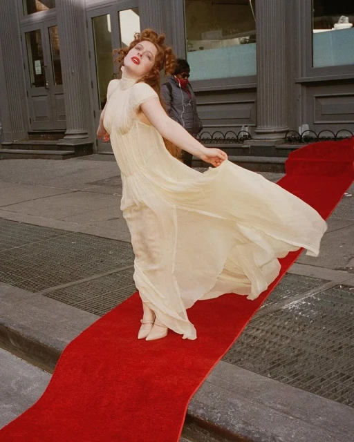A woman in a long cream dress poses on a red carpet outdoors with a historic-style building featuring columns in the background.