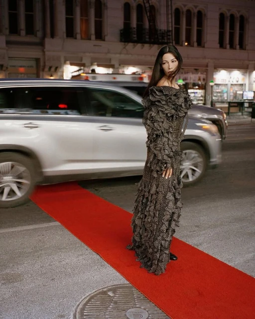 Woman in a ruffled gown standing on a red carpet outdoors at night, with a blurry car in the background and city buildings.