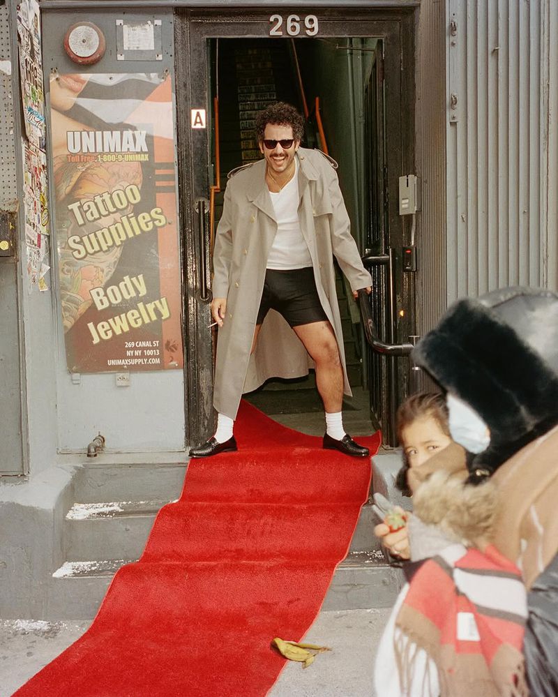 A man in a trench coat stands in a doorway on a red carpet, smiling. A tattoo and body jewelry store poster is visible in the background.