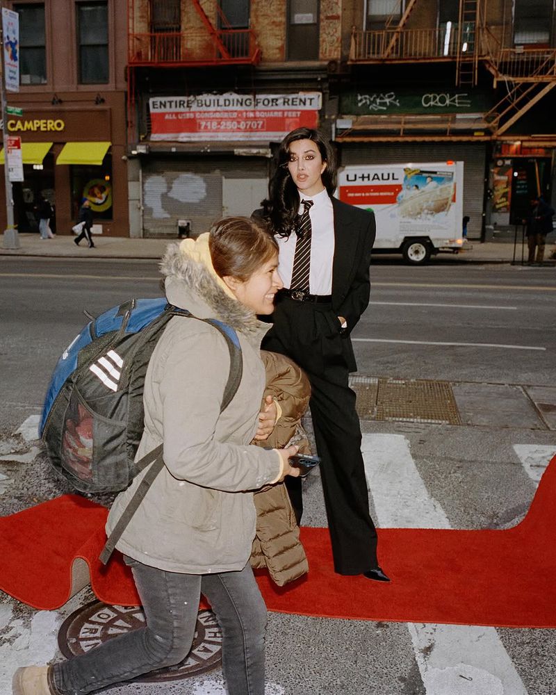 Elegant woman in a suit stands on a red carpet in an urban setting, with a pedestrian walking past, showcasing fashion and street style.