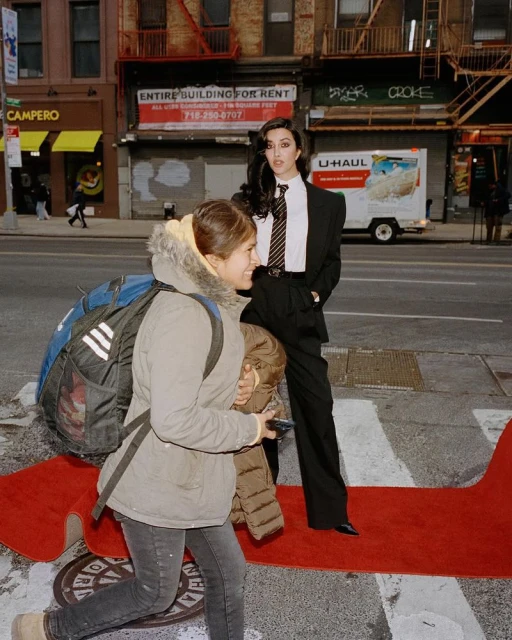 Elegant woman in a suit stands on a red carpet in an urban setting, with a pedestrian walking past, showcasing fashion and street style.