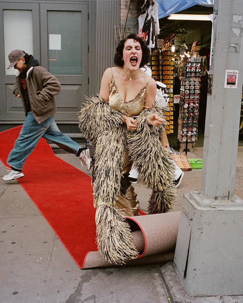 Woman in a gold dress and feathered scarf poses on red carpet. Man walks by in background on urban sidewalk. Bright daylight.