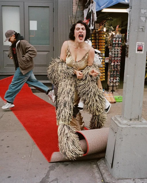 Woman in a gold dress and feathered scarf poses on red carpet. Man walks by in background on urban sidewalk. Bright daylight.