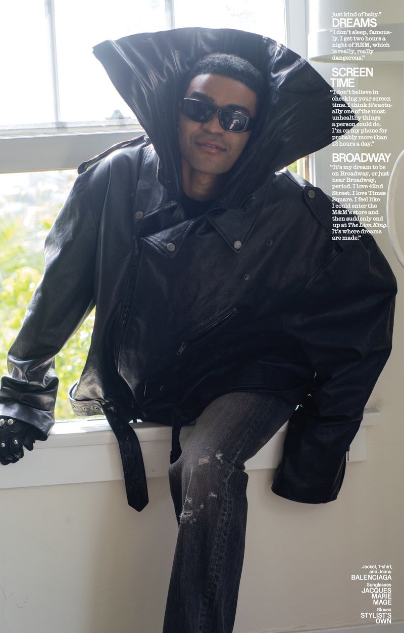 Man in oversized black leather jacket, stylish sunglasses, seated on window sill with muted background, editorial fashion shot.