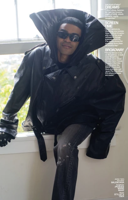 Man in oversized black leather jacket, stylish sunglasses, seated on window sill with muted background, editorial fashion shot.
