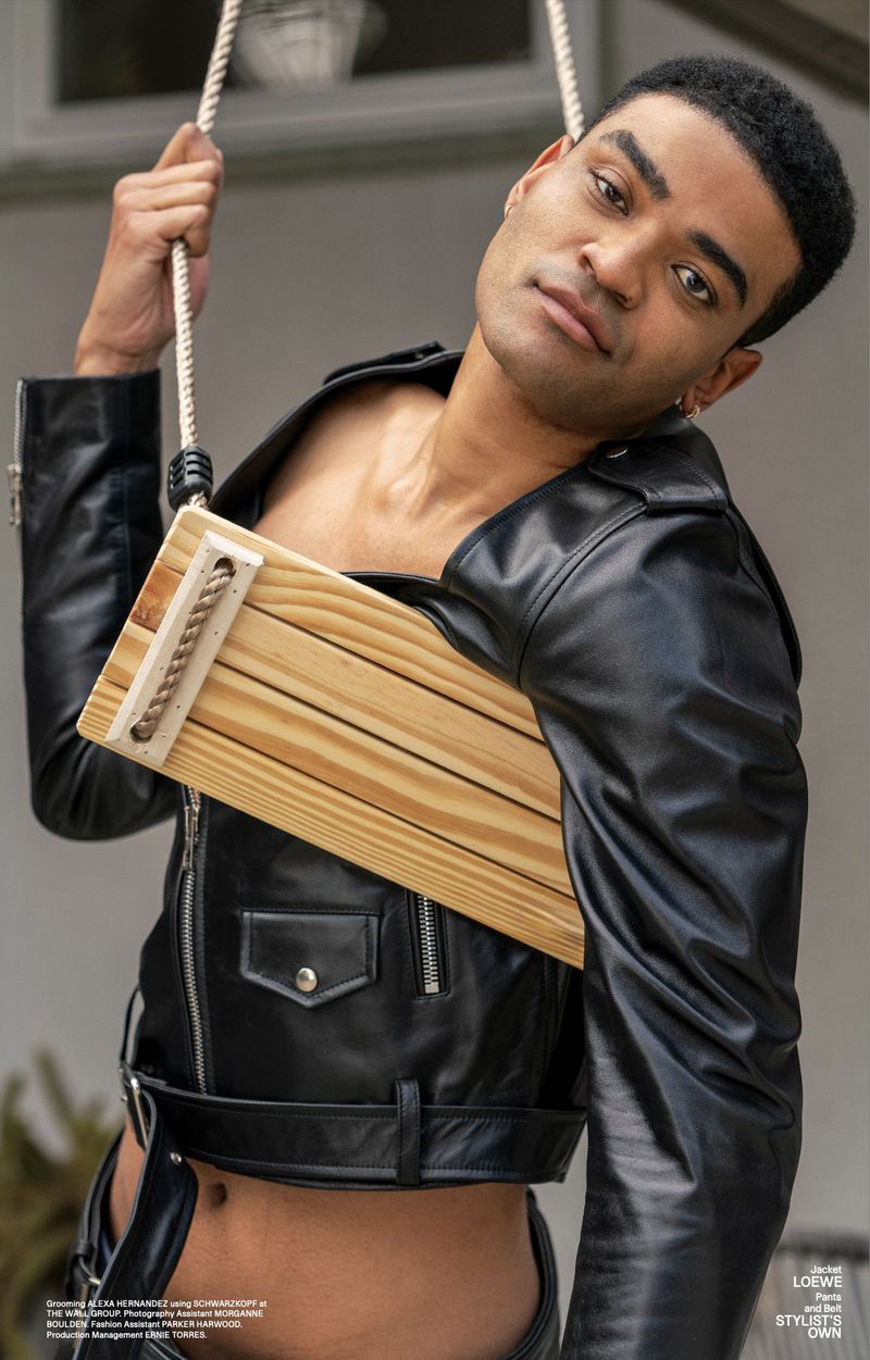 Editorial shot of a light-skinned male model in a black leather jacket and trousers, posing with a wood prop swing indoors.