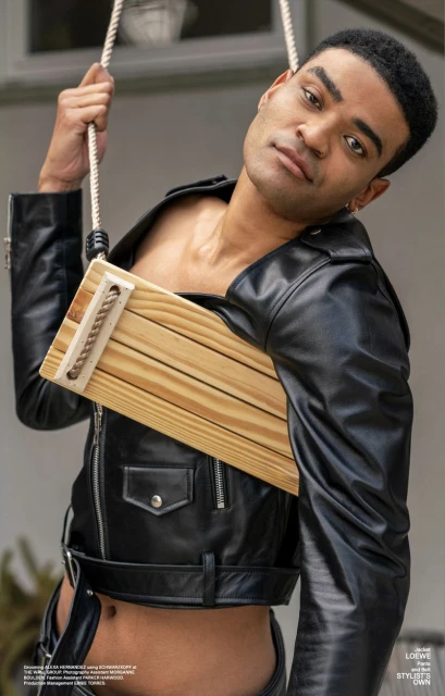 Editorial shot of a light-skinned male model in a black leather jacket and trousers, posing with a wood prop swing indoors.