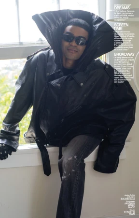 Man wearing an oversized black leather jacket with a high collar and sunglasses seated near a window for Interview Magazine