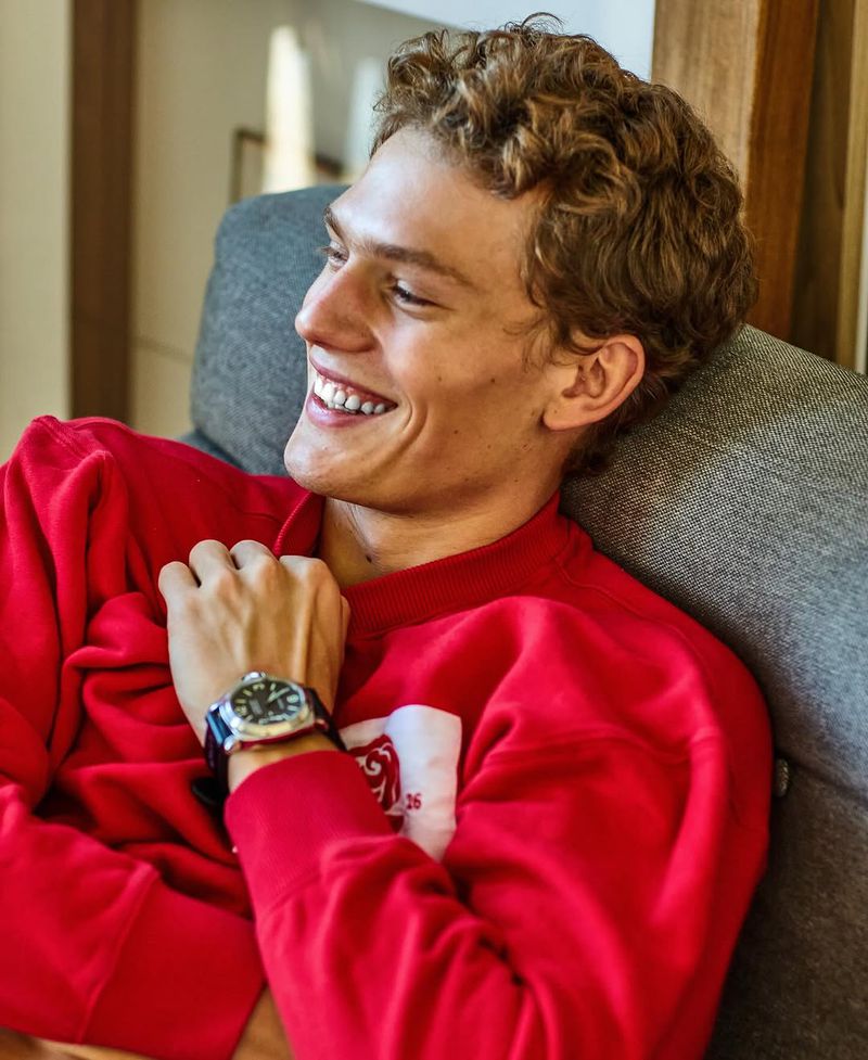 Smiling young adult male with curly hair, wearing a red sweatshirt and a watch, is relaxing on a gray couch indoors in soft light.