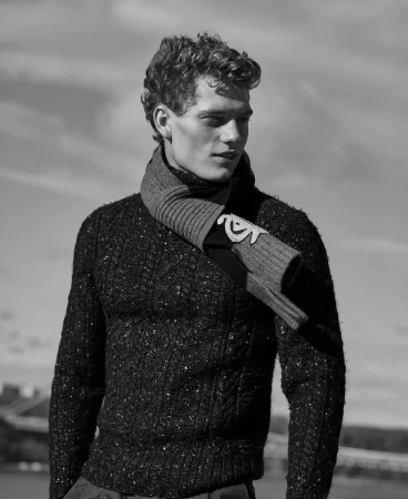 Black and white portrait of a handsome adult male model with curly hair wearing a sweater and scarf with a blurred building background.