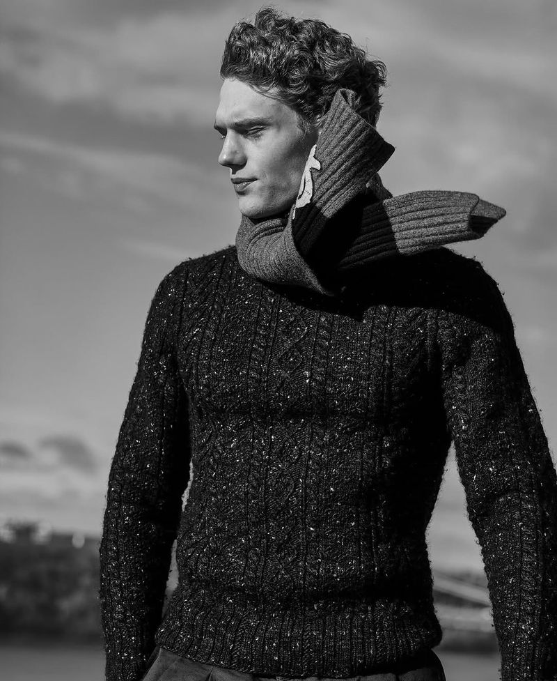 Monochrome portrait of a male model wearing a wool sweater and scarf against a sky background, fashion editorial style.