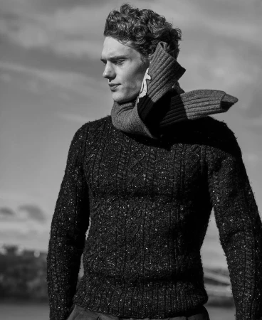 Monochrome portrait of a male model wearing a wool sweater and scarf against a sky background, fashion editorial style.