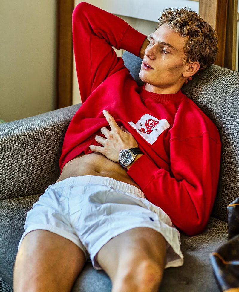 Young adult man lounges on a gray sofa wearing a red sweatshirt, white shorts, and a wristwatch in a casual indoor setting.