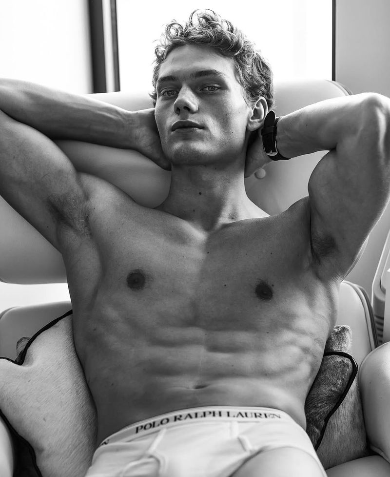 Black and white portrait of a muscular adult male with curly hair, arms raised behind his head, sitting in a chair wearing underwear.