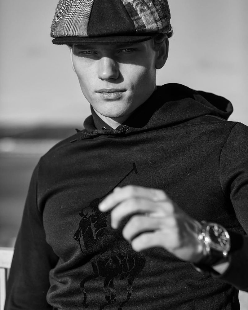 Monochrome portrait of a young man wearing a newsboy cap and hoodie with a logo, captured in soft outdoor lighting.