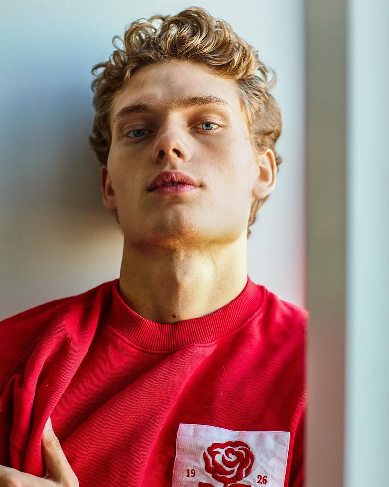 Close-up portrait of a young adult male with curly hair wearing a red sweatshirt, posing against a light background in a studio setting.