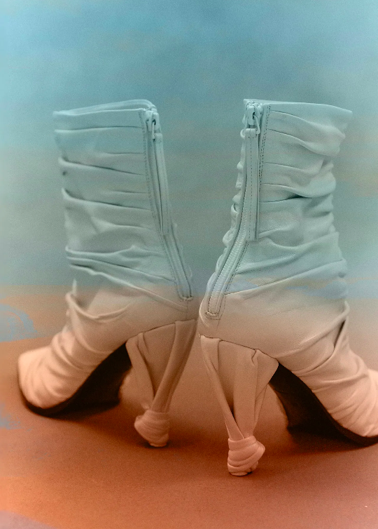 Two light blue heeled boots with zippers are presented on a neutral surface. The backdrop is a soft gradient of blues and yellows.