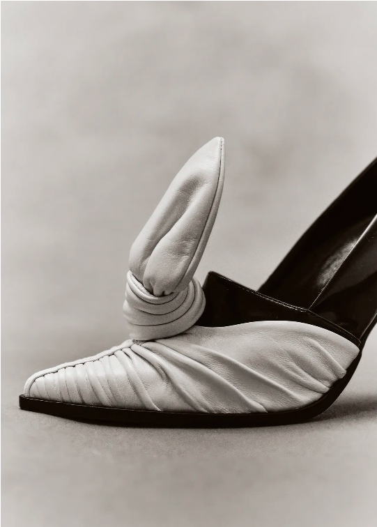 Monochromatic still life showcasing a white leather high heel shoe with a unique ruched design and decorative knot against a neutral background.