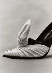 Close-up of a white and black pointed-toe high heel shoe with sculptural knotted leather detailing on a neutral background.